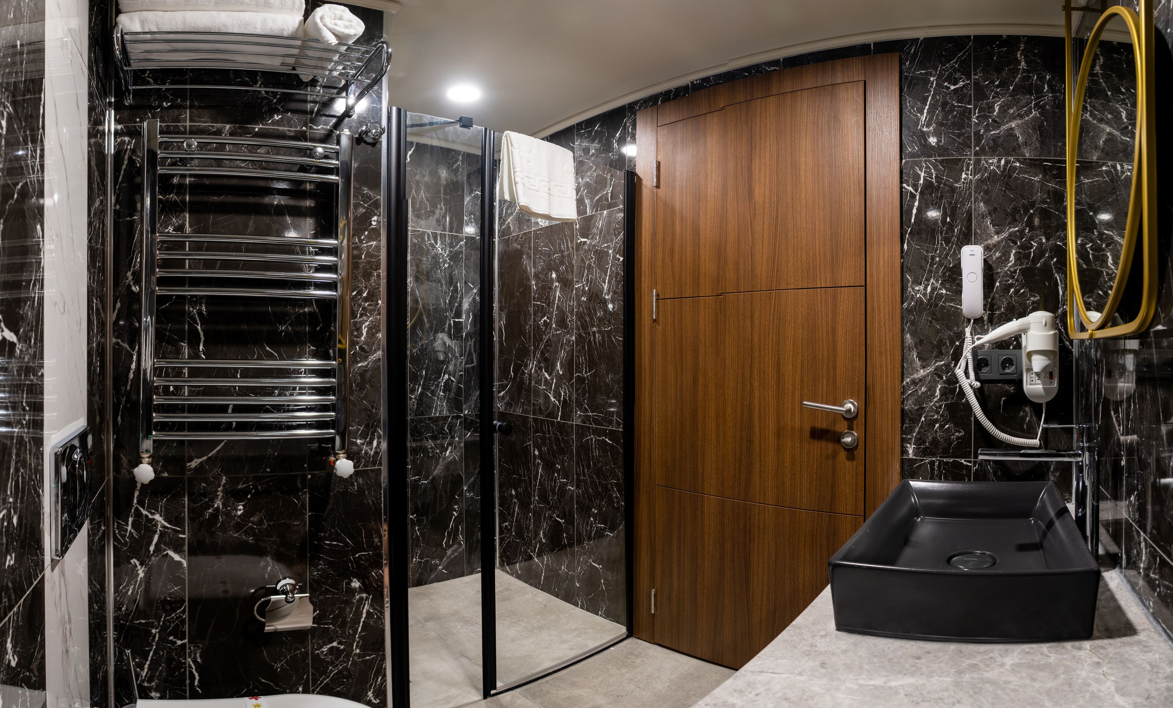 Un baño moderno con cabina de ducha de vidrio, puerta de madera, lavabo negro y pared con azulejos de mármol.