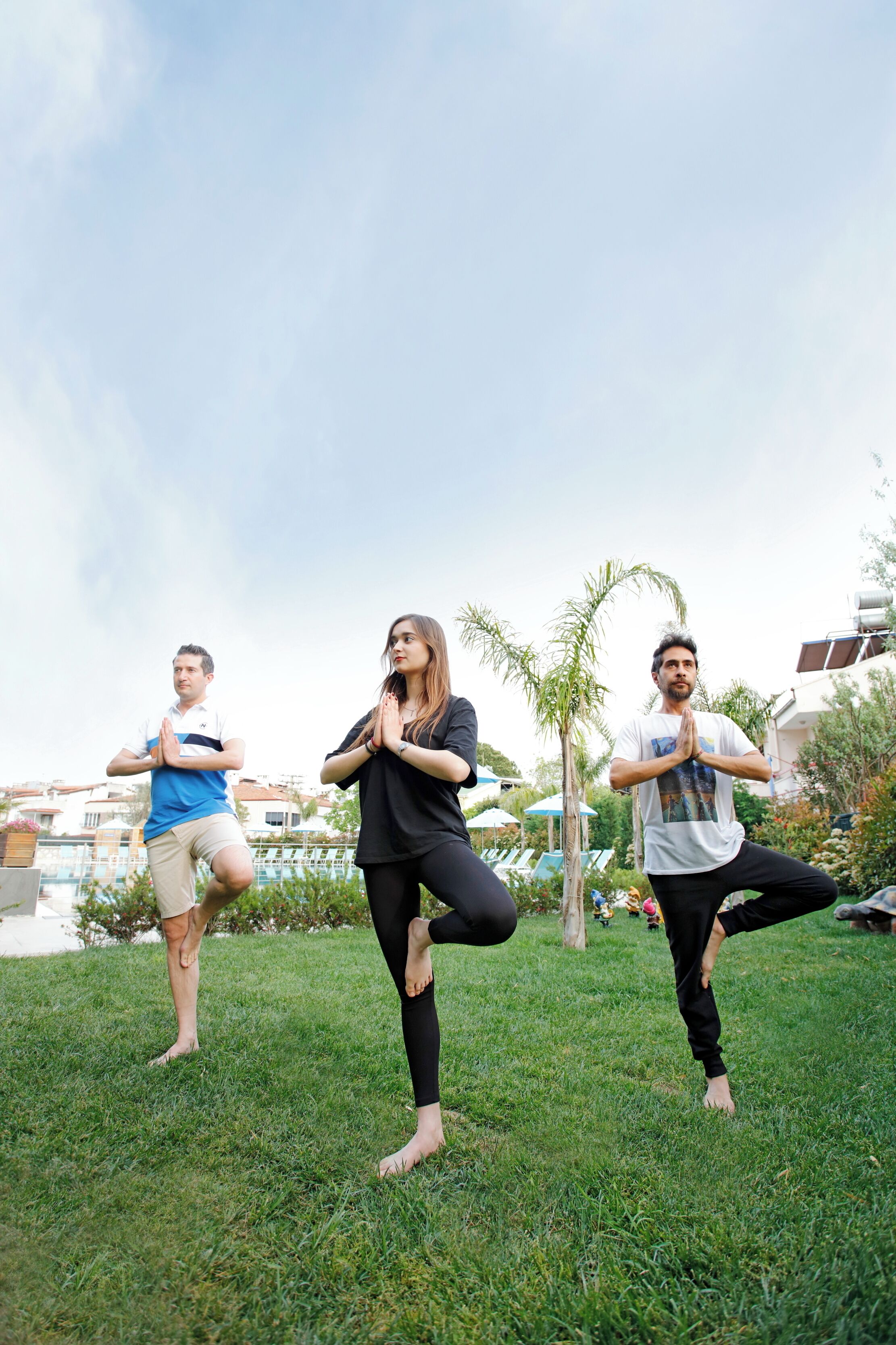 Three people performing yoga in an outdoor grassy area with buildings in the background.