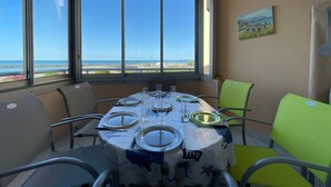 Een eetruimte met een tafel voor vier personen, grote ramen met uitzicht op zee en een schilderij aan de muur.