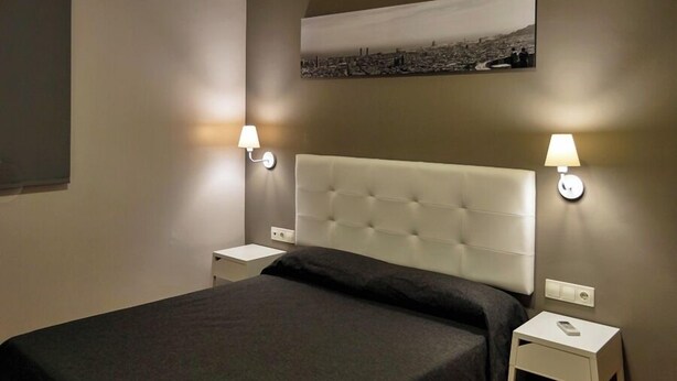 A neatly made bed with a dark bedspread, two bedside tables with lamps, and a cityscape artwork on the wall. - Hostal Sant Pau (Barcelona)