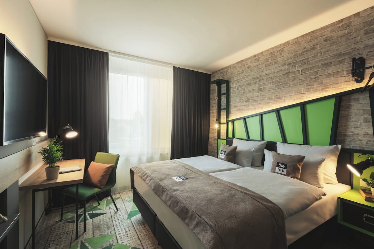 A modern hotel room with a large bed, a desk with a chair, a television, and a brick accent wall.