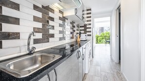 Et moderne køkken med en sort bordplade, stålvask og et bagpanel med en blanding af hvide og brune fliser. Der er et vindue over vasken, og der er udsigt til en balkon med gelænder.