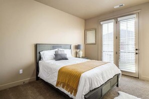 Un dormitorio con cama, mesita de noche, lámpara, puerta con persianas y un espejo adosado a la pared.