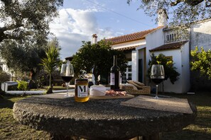 A stone table with a bottle of olive oil, a glass of red wine, and a plate of cured meats in a garden setting. - A Casa da Ti Vina - Casa Típica Alentejana (Gavião)