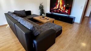 A modern living room with a sectional sofa, a wooden coffee table, and a flat-screen TV displaying a fire. - Ferienwohnung am Jacobsweg (Wurzen)