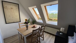 A dining area with a table, chairs, a vase with flowers, and a plate of fruit. There are two windows providing natural light and a view of greenery outside. - Ferienwohnung am Jacobsweg (Wurzen)