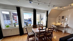 A dining area with a wooden table and chairs, large windows with curtains, a radiator, and a desk with a lamp. - Ferienwohnung am Jacobsweg (Wurzen)
