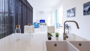 Cozinha moderna com bancada branca, duas pias e torneira de aço inoxidável. Há duas plantas em vasos sobre a bancada e uma janela grande com persianas à esquerda.