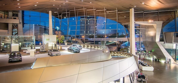 A modern building with a curved glass facade, multiple floors, and a central atrium with cars displayed. - Holiday Inn - the niu, Brass Munich Olympiapark by IHG (Munich)