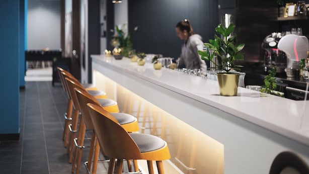 A modern bar area with wooden bar stools, a long counter, and potted plants. - Holiday Inn - the niu, Brass Munich Olympiapark by IHG (Munich)