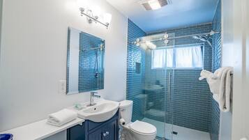 A bathroom with a sink, mirror, and toilet. There is a glass shower enclosure with a window.