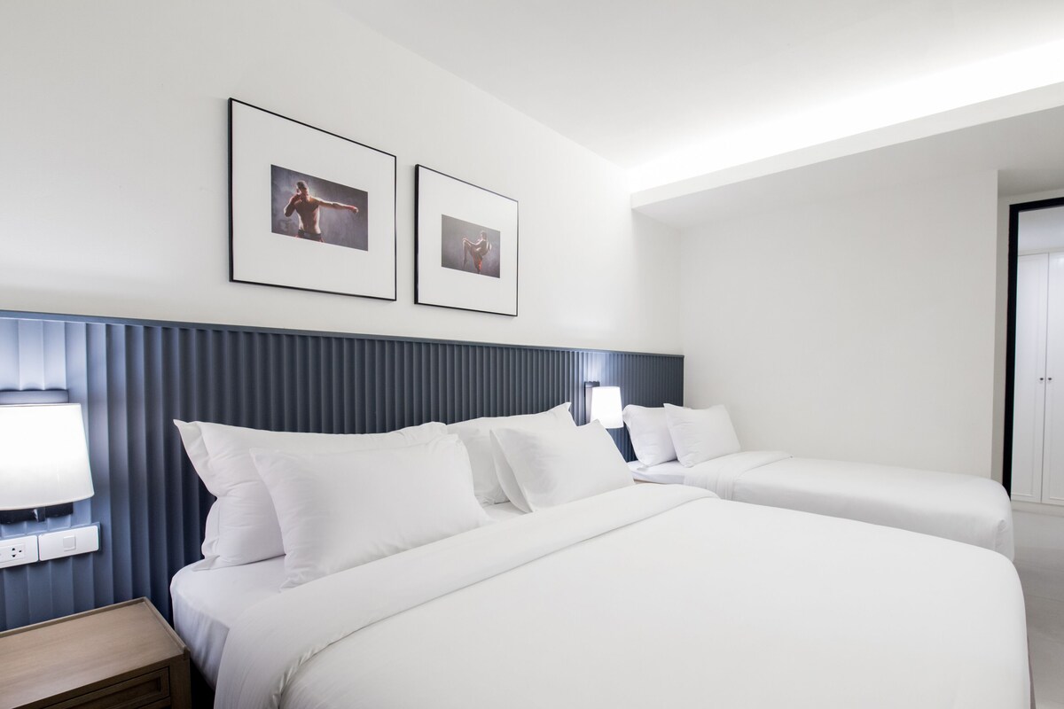 Habitación de hotel con dos camas, una mesita de noche con lámpara, y dos cuadros en la pared.