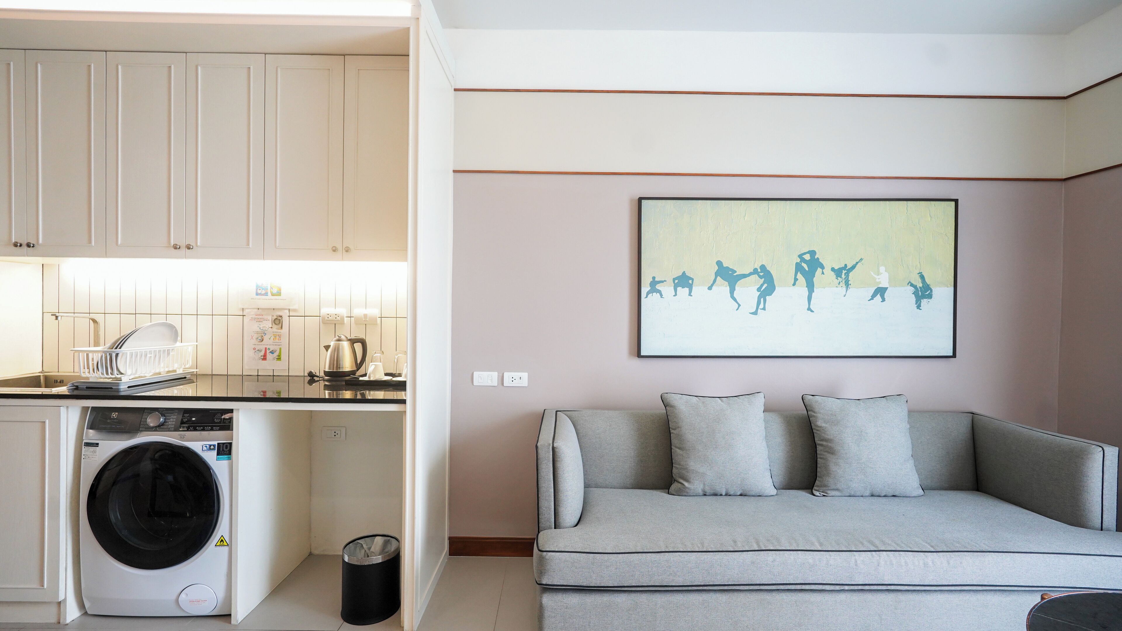Un espace de vie compact comprenant un coin cuisine, un lave-linge, un canapé et une œuvre d’art accrochée au mur.