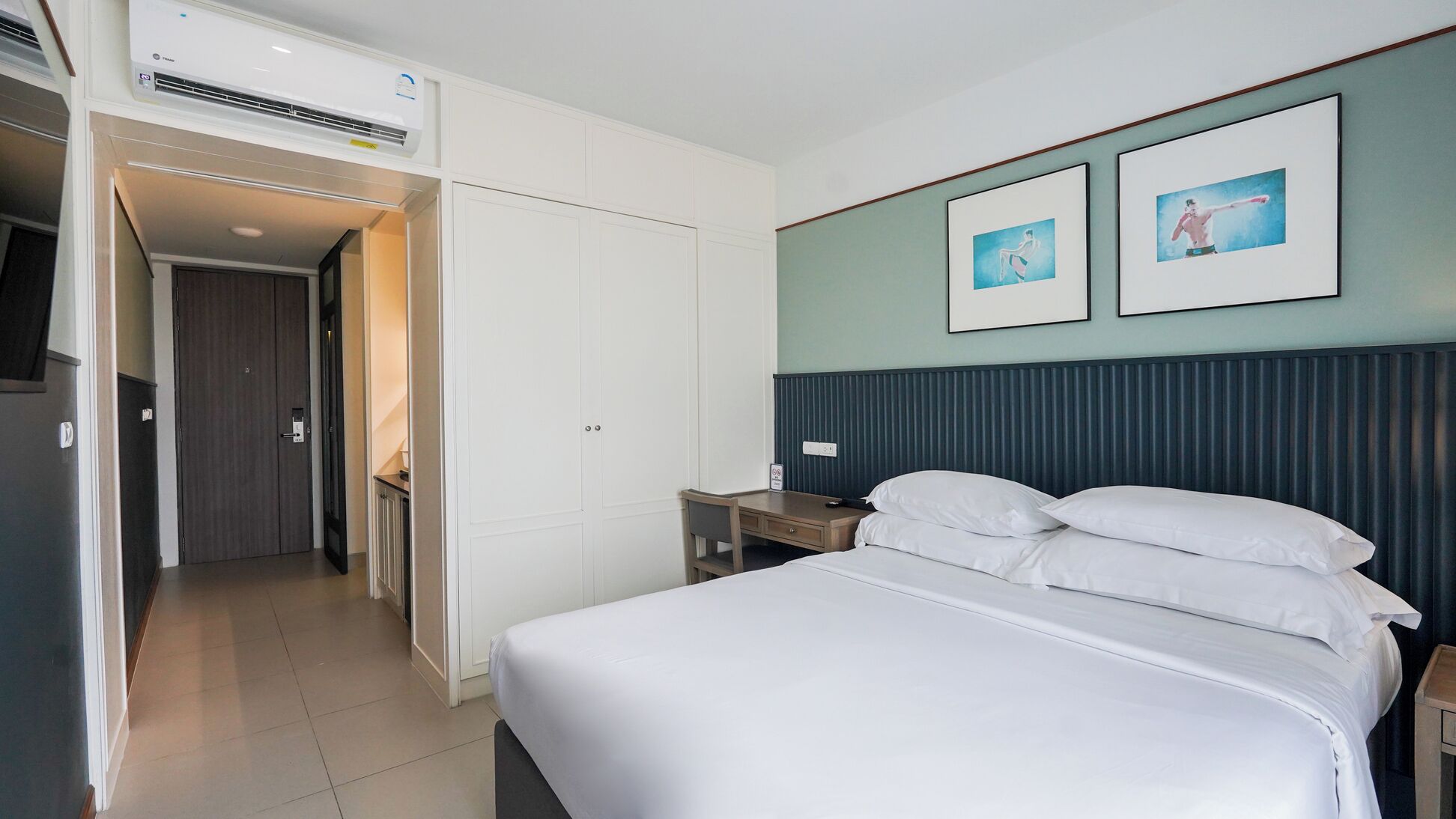 Ein Hotelzimmer mit Bett, Nachttisch und Schreibtisch mit Stuhl. Außerdem ist ein Kleiderschrank und eine Klimaanlage vorhanden.
