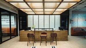 Un área de recepción con un escritorio de madera, tres taburetes de bar, una planta en maceta y una cita en la pared de vidrio.