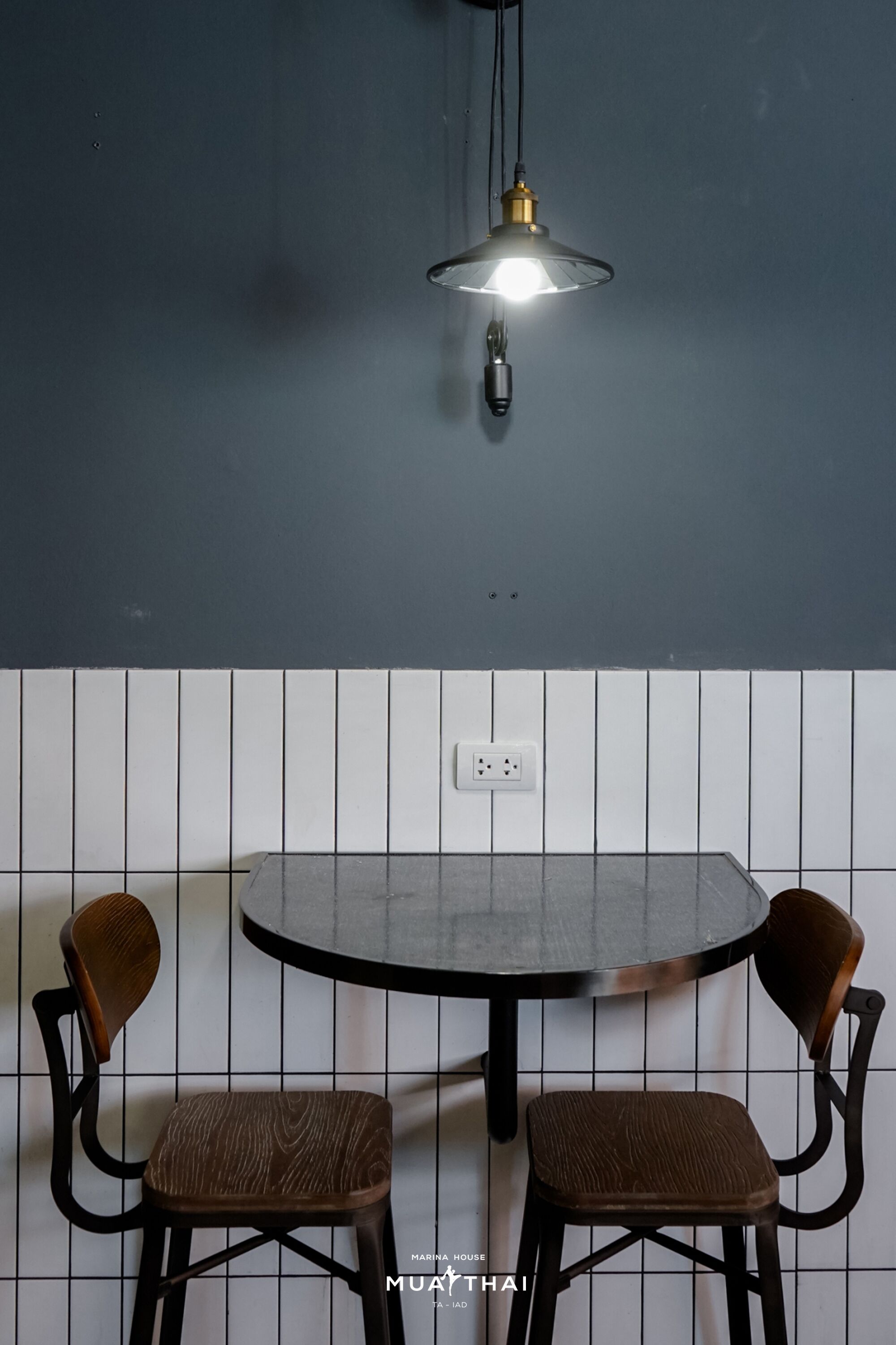 A dining area with a round table and two chairs, a pendant light, and a tiled wall with a power outlet.