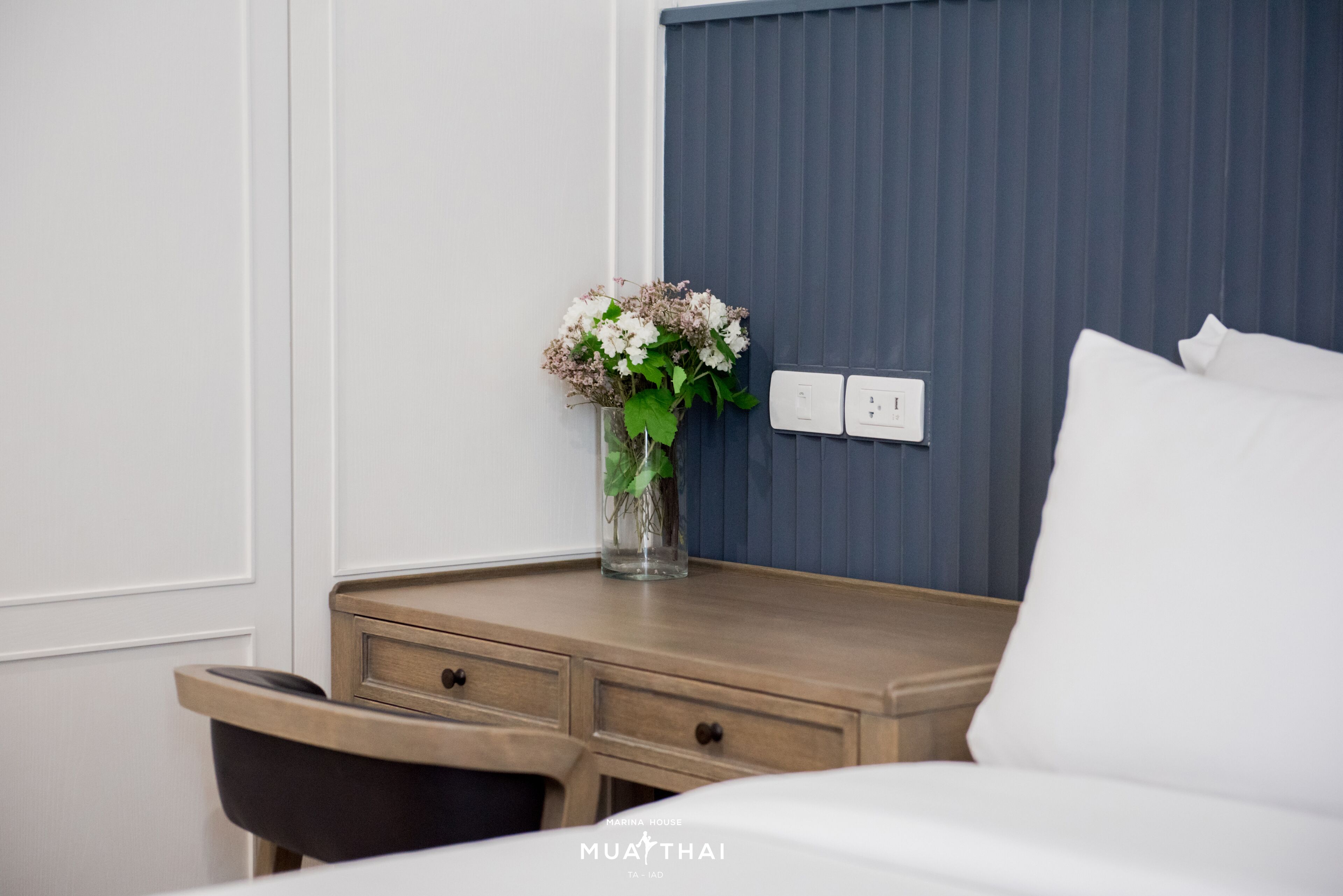 A hotel room with a wooden nightstand, a vase with flowers, a bed with white bedding, and a blue headboard.