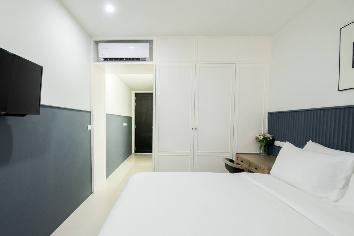 A modern hotel room with a large bed, a flat-screen TV mounted on the wall, a wardrobe, and a bedside table with a vase of flowers.