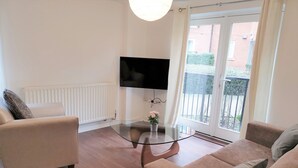 Un salon moderne avec une table basse en verre, un canapé beige, un téléviseur à écran plat fixé au mur et une vue sur un bâtiment en briques, visible à travers une porte vitrée.