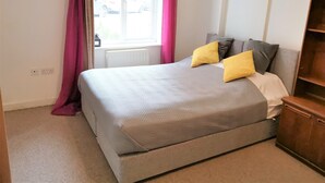 Une chambre à coucher avec un lit, une fenêtre avec des rideaux, un radiateur et une table de chevet en bois.