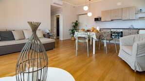 A modern living room with a sofa, dining table, and chairs. There is a kitchen area with cabinets and a microwave. - Casa Baia Blu (San Lorenzo al Mare)