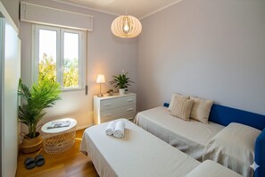 A bedroom with two beds, a bedside table, a lamp, a potted plant, and a window with blinds.