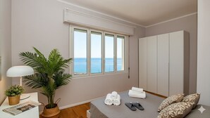 A bedroom with a large window, a bed with pillows and towels, a bedside table with a lamp and a plant, and a wardrobe. - Casa Baia Blu (San Lorenzo al Mare)