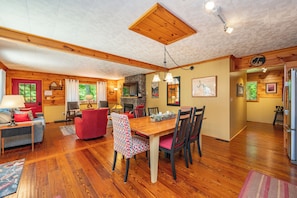 A cozy cabin interior with a wooden dining table, chairs, a fireplace, and a kitchen area.