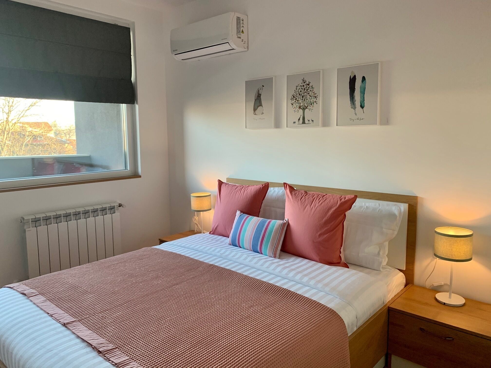 A neatly made bed with a pink and white striped blanket, two pink pillows, and a striped pillow. There are two bedside lamps, a window with a black blind, and an air conditioning unit above the bed.