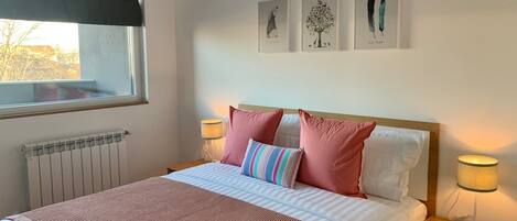 A neatly made bed with a pink and white striped blanket, two pink pillows, and a striped pillow. There are two bedside lamps, a window with a black blind, and an air conditioning unit above the bed.