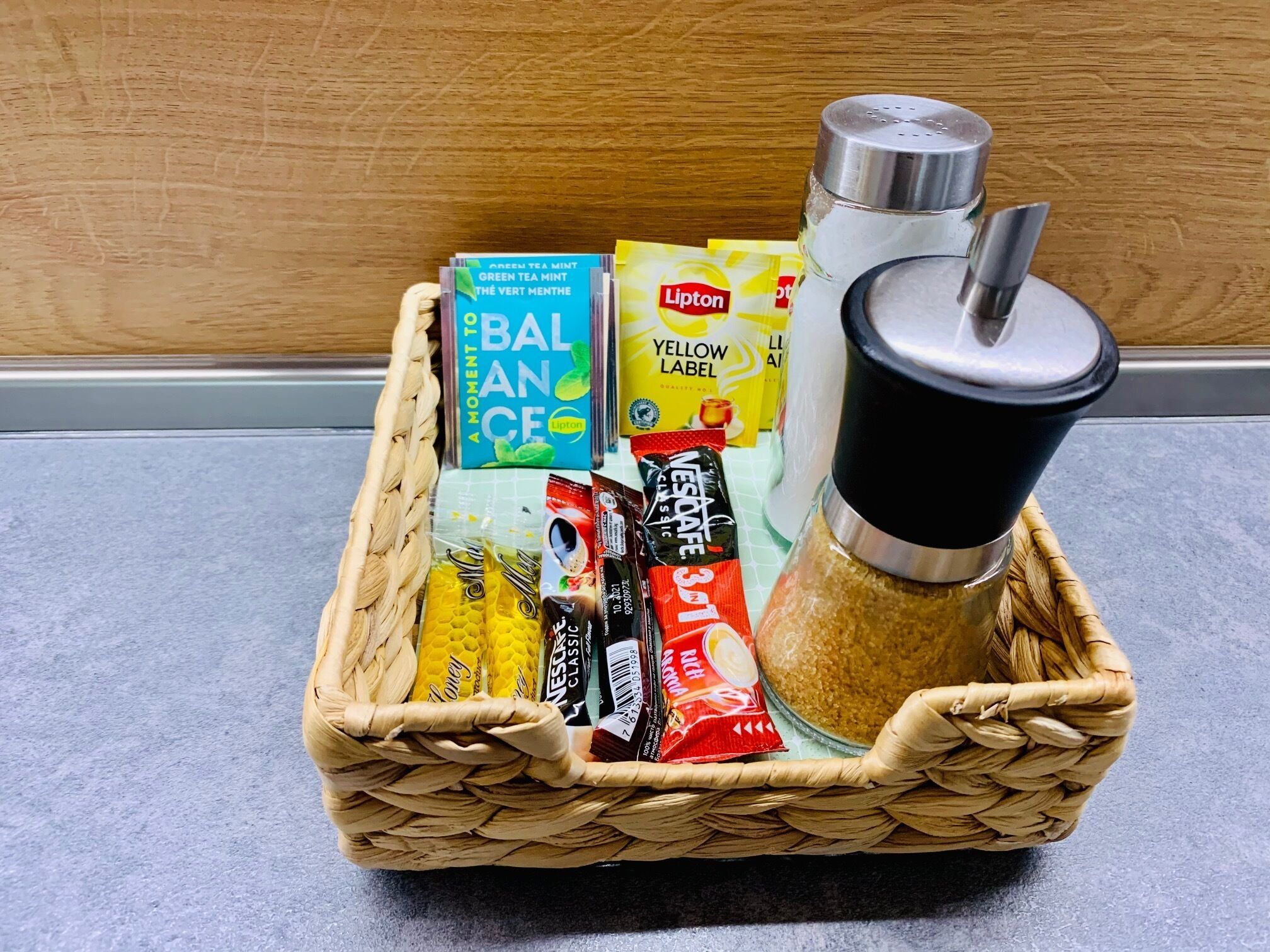 A woven basket containing a variety of packaged goods, including a bottle of Lipton tea, a box of Nescafé Classic, and a yellow label.