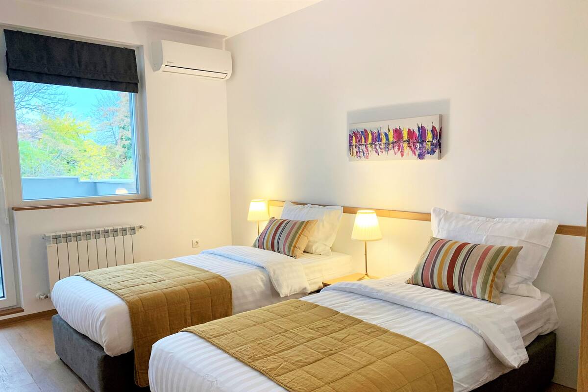 Une chambre d’hôtel avec deux lits, un radiateur, un climatiseur, une fenêtre donnant sur des arbres et une œuvre d’art colorée accrochée au mur.