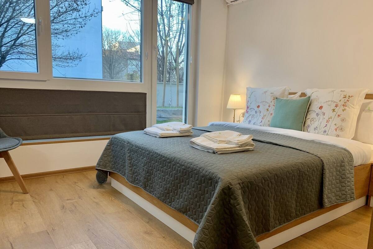 Un lit bien fait, recouvert d’une courtepointe matelassée, une chaise et une fenêtre donnant sur des arbres et des immeubles.