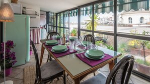 Zona de menjador amb una taula preparada per a quatre, grans finestres amb vista a la ciutat i una cuineta al fons.