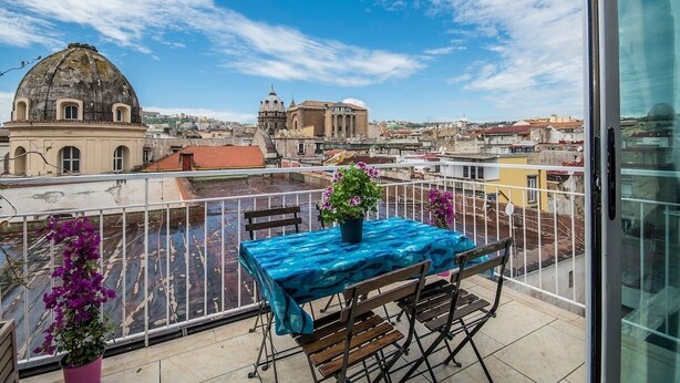 Un balcó amb taula i cadires, amb vista a un paisatge urbà ple d'edificis històrics i un cel serè.