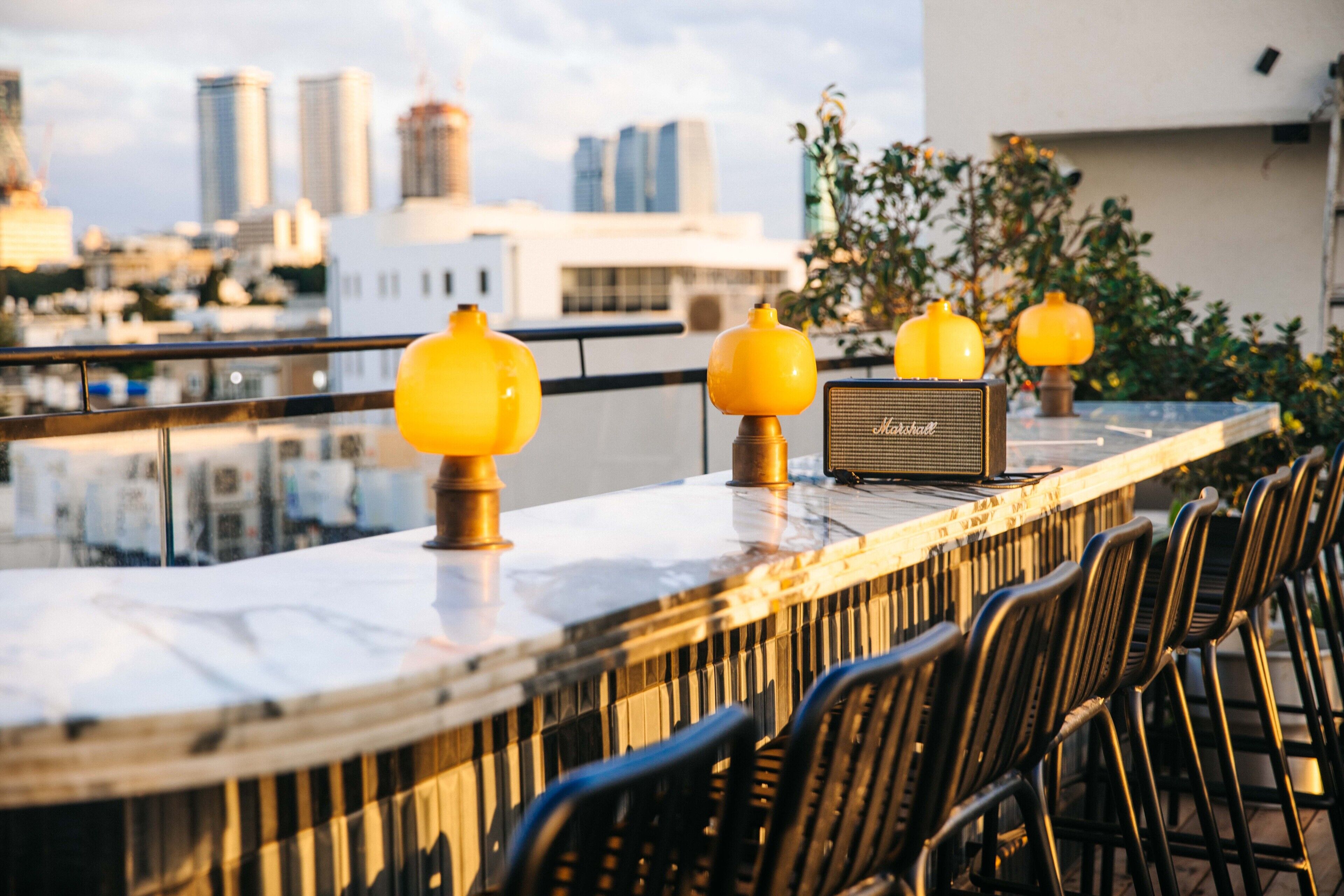 A rooftop bar with a city view, featuring a marble counter, yellow spherical lights, and black chairs.