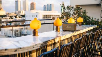Un bar situé sur le toit d’un immeuble, offrant une vue sur la ville, avec un comptoir en marbre, des lampes sphériques jaunes et des chaises noires.