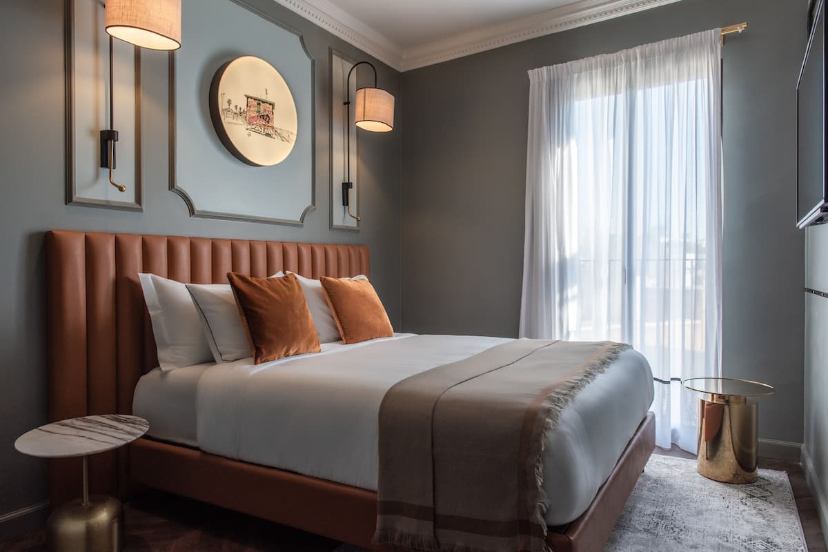 Un dormitorio moderno con una cama grande, un espejo redondo, dos lámparas de pared y vistas a un paisaje urbano a través de cortinas translúcidas.