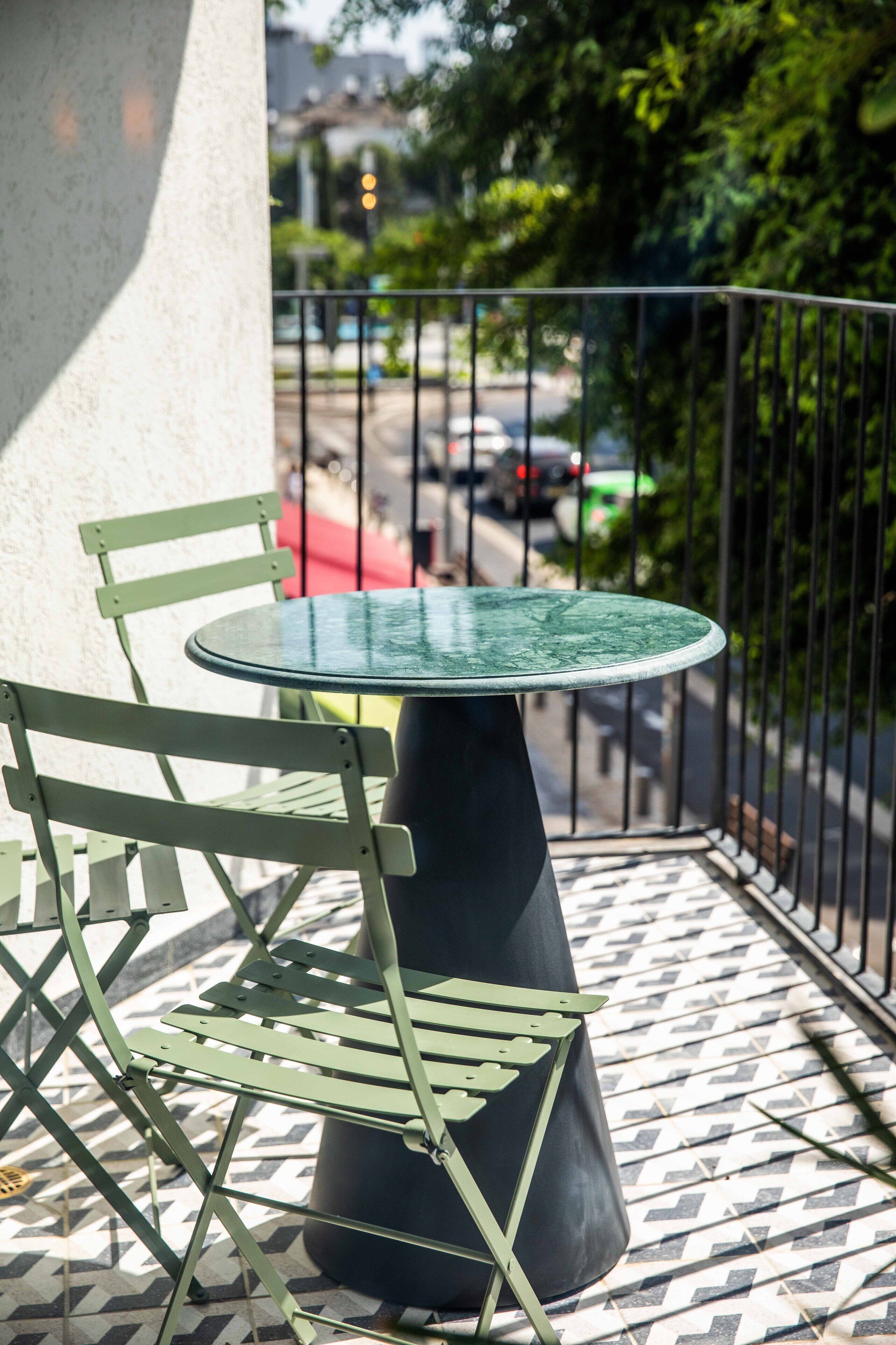 A small outdoor seating area with a round table and two chairs, a patterned floor, and a view of a street with cars and greenery.