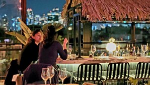 Un área de comedor al aire libre con techo de paja, mesas dispuestas con copas de vino y vistas a la ciudad de fondo.