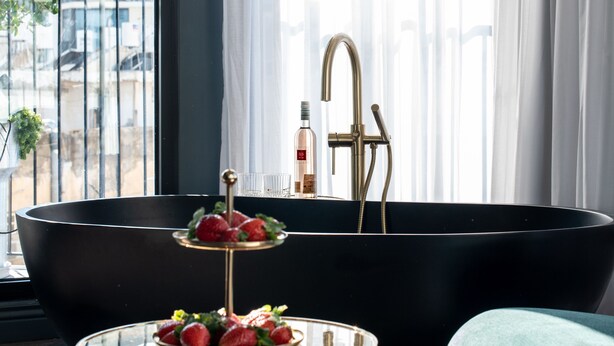 Un baño moderno con una bañera negra, un grifo dorado y una copa de vino en el alféizar.