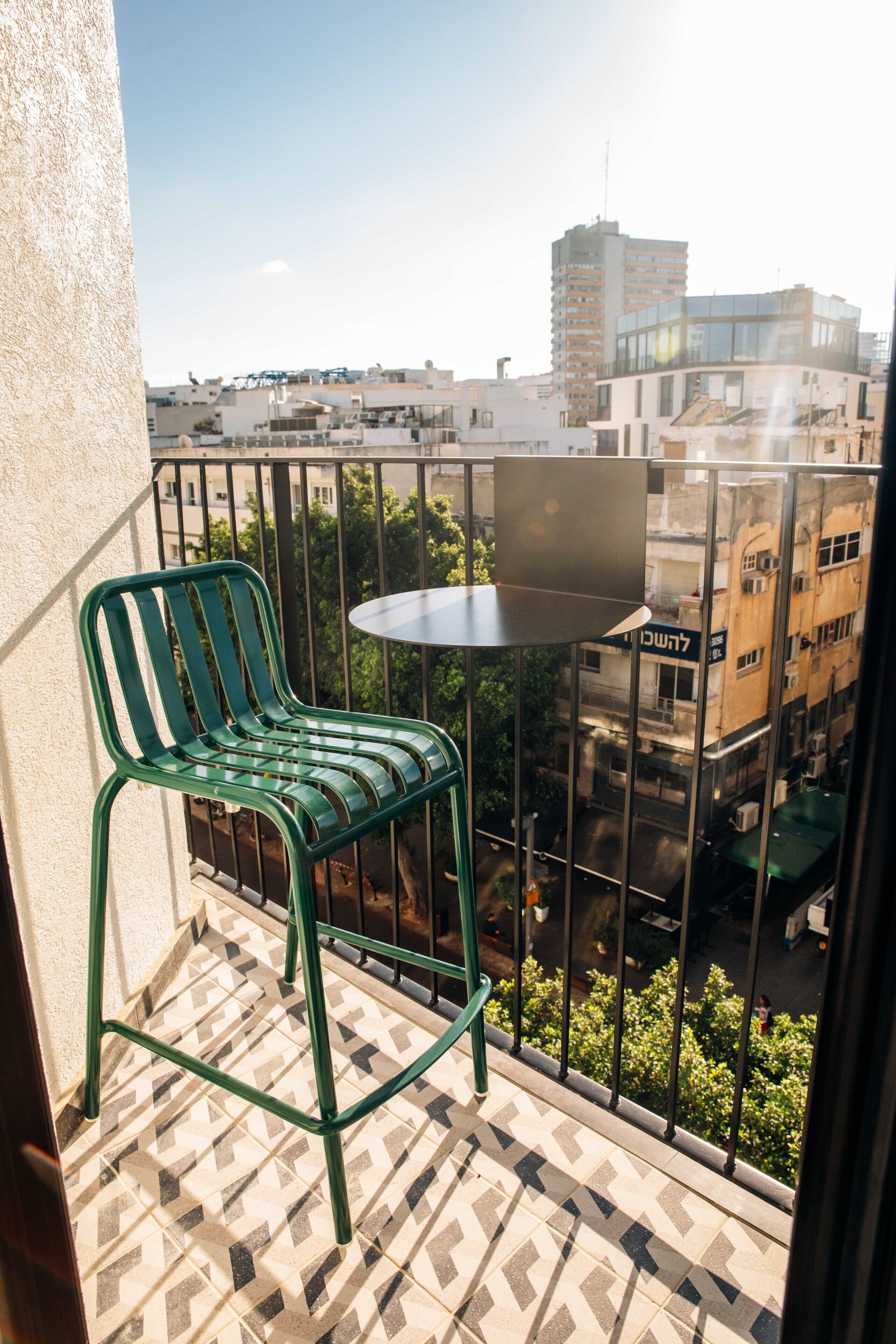 A balcony with a green chair and a small round table, overlooking a cityscape with buildings and trees.