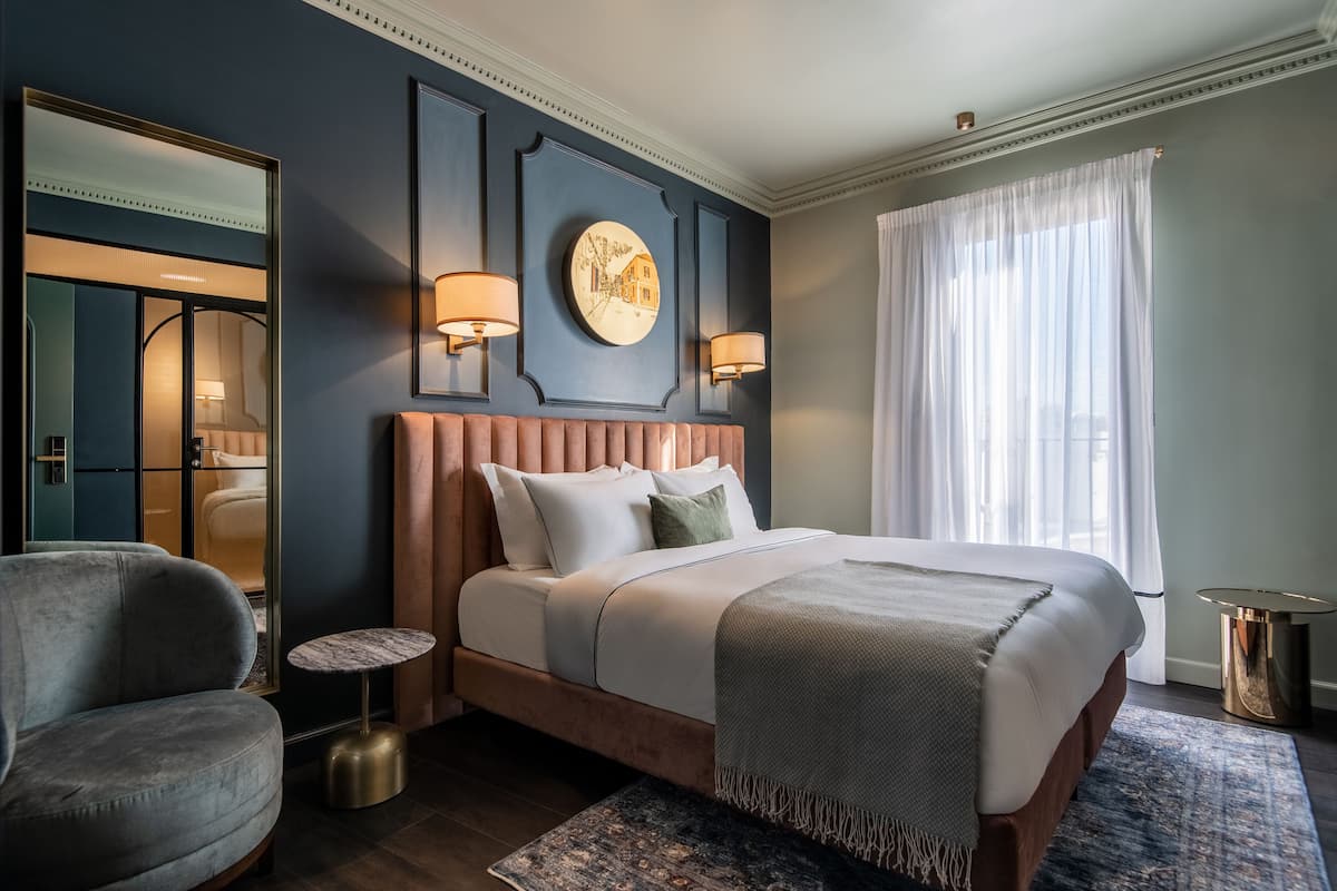 Una habitación de hotel moderna con una cama grande, un reloj de pared redondo, dos lámparas fijadas a la pared y un ventanal con cortinas.