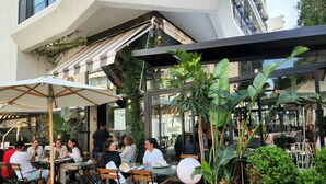 Un café al aire libre con mesas y sillas, rodeado de plantas en macetas y sombrillas.