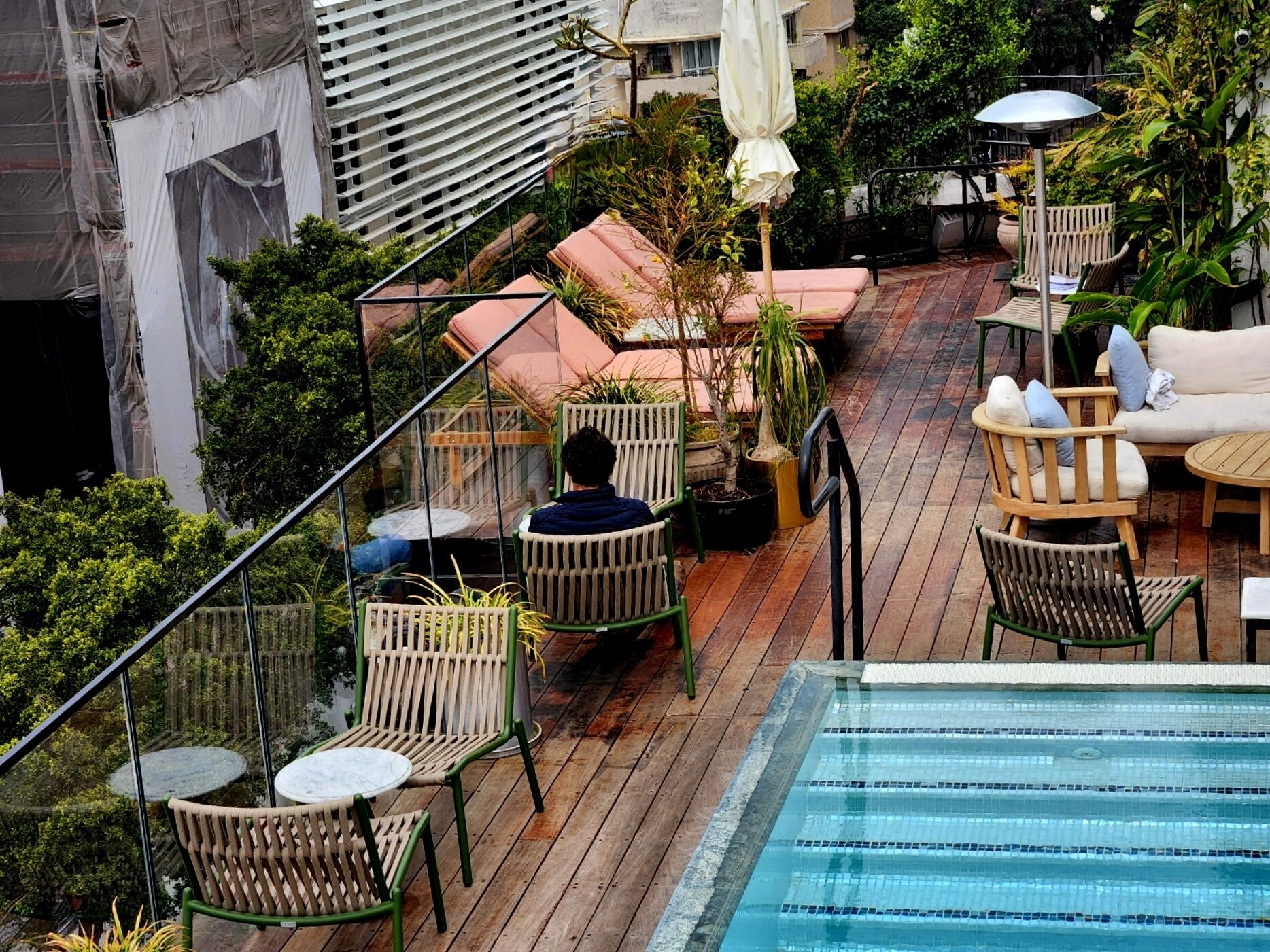 Un área de piscina en la azotea con cubierta de madera, sillas de descanso y barandilla de cristal con vistas a un paisaje urbano.