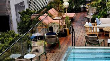Un área de piscina en la azotea con cubierta de madera, sillas de descanso y barandilla de cristal con vistas a un paisaje urbano.