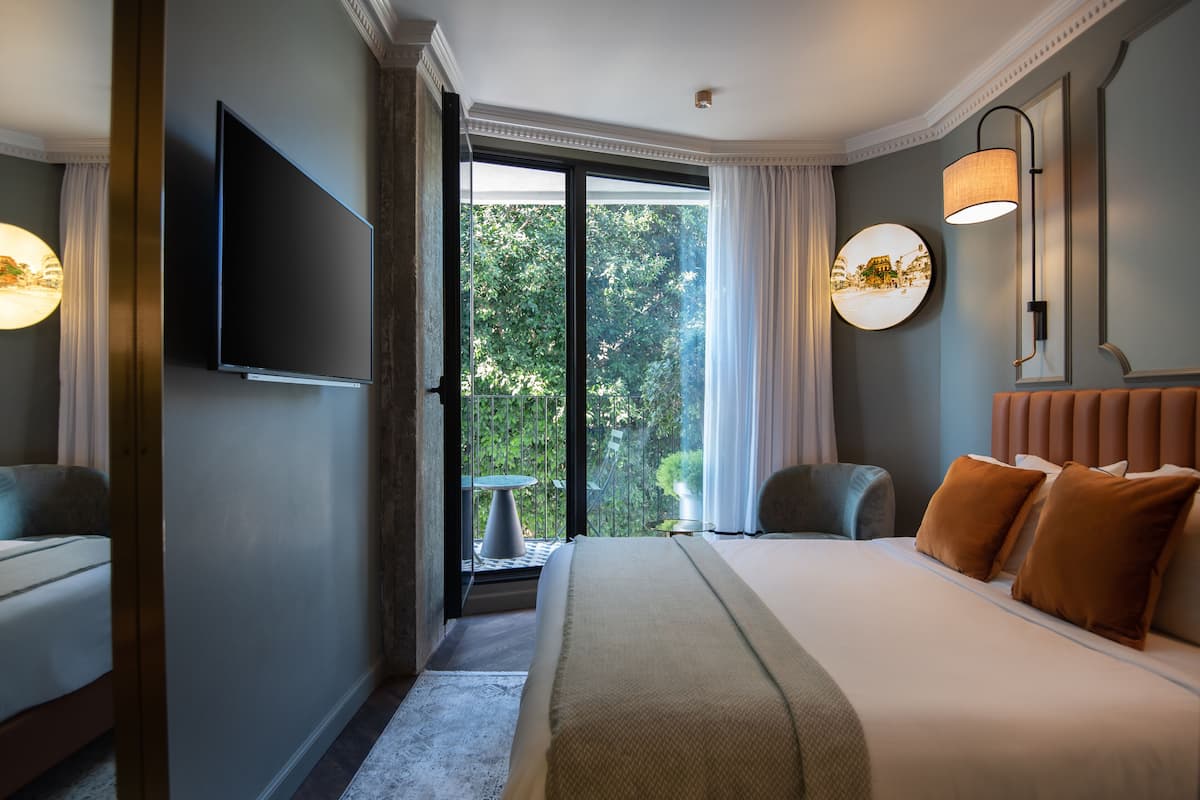 Habitación de hotel con una cama grande, un televisor de pantalla plana, un balcón con mesa y sillas, y vista a zonas verdes.