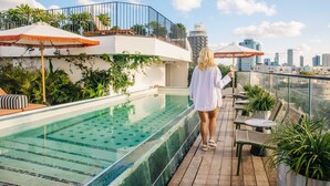 Un área de piscina en la azotea con vista al horizonte de la ciudad, un edificio blanco con balcón y varias plantas en macetas.