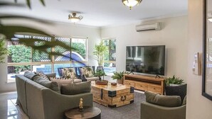 Een moderne woonkamer met een zithoek, een salontafel en een televisie. Er staan planten en een groot raam met uitzicht op groen.