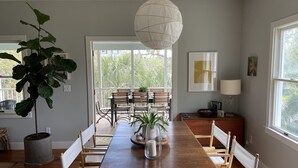 A dining area with a wooden table, chairs, a hanging light fixture, and a large window with a view of greenery. - Fun Beach House (Sullivans Island)